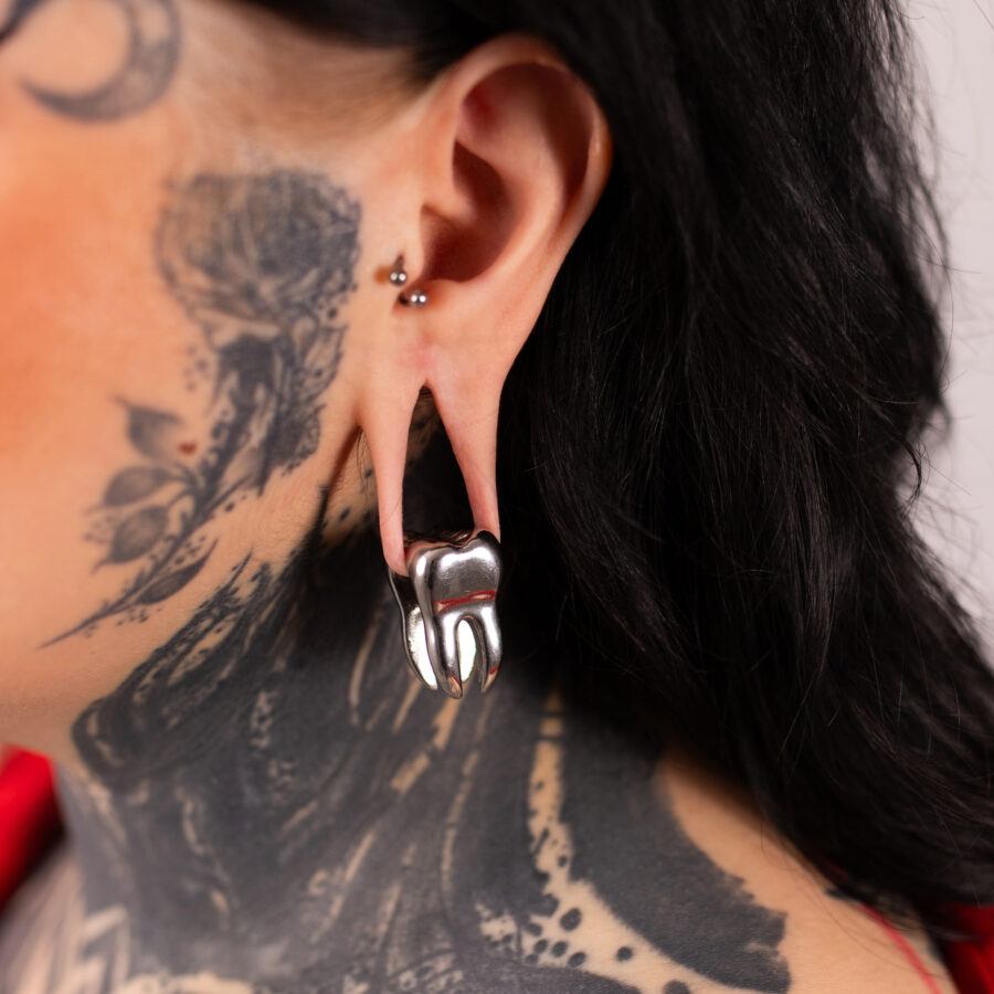 Peso de oreja plateado en forma de diente en la oreja de una persona tatuada con cabello negro.
