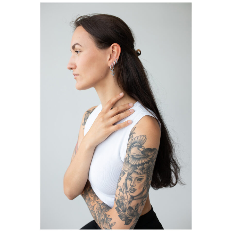 Mujer con cabello largo y brazos tatuados, lleva pendientes de plata Little Anchor Hoops