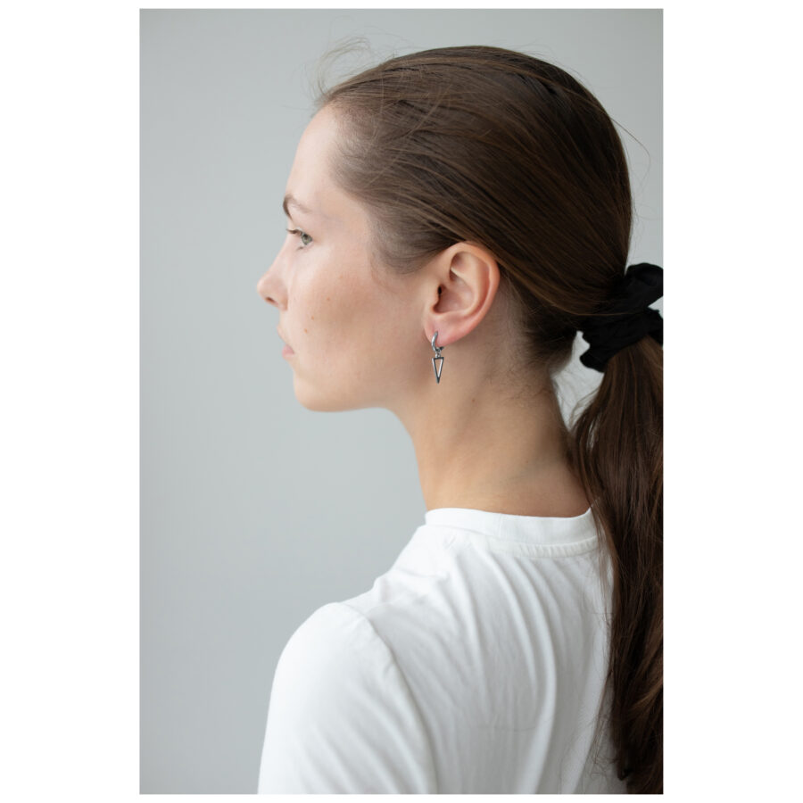 Mujer con coleta lateral lleva pequeños aros triangulares negros y plateados sobre fondo gris
