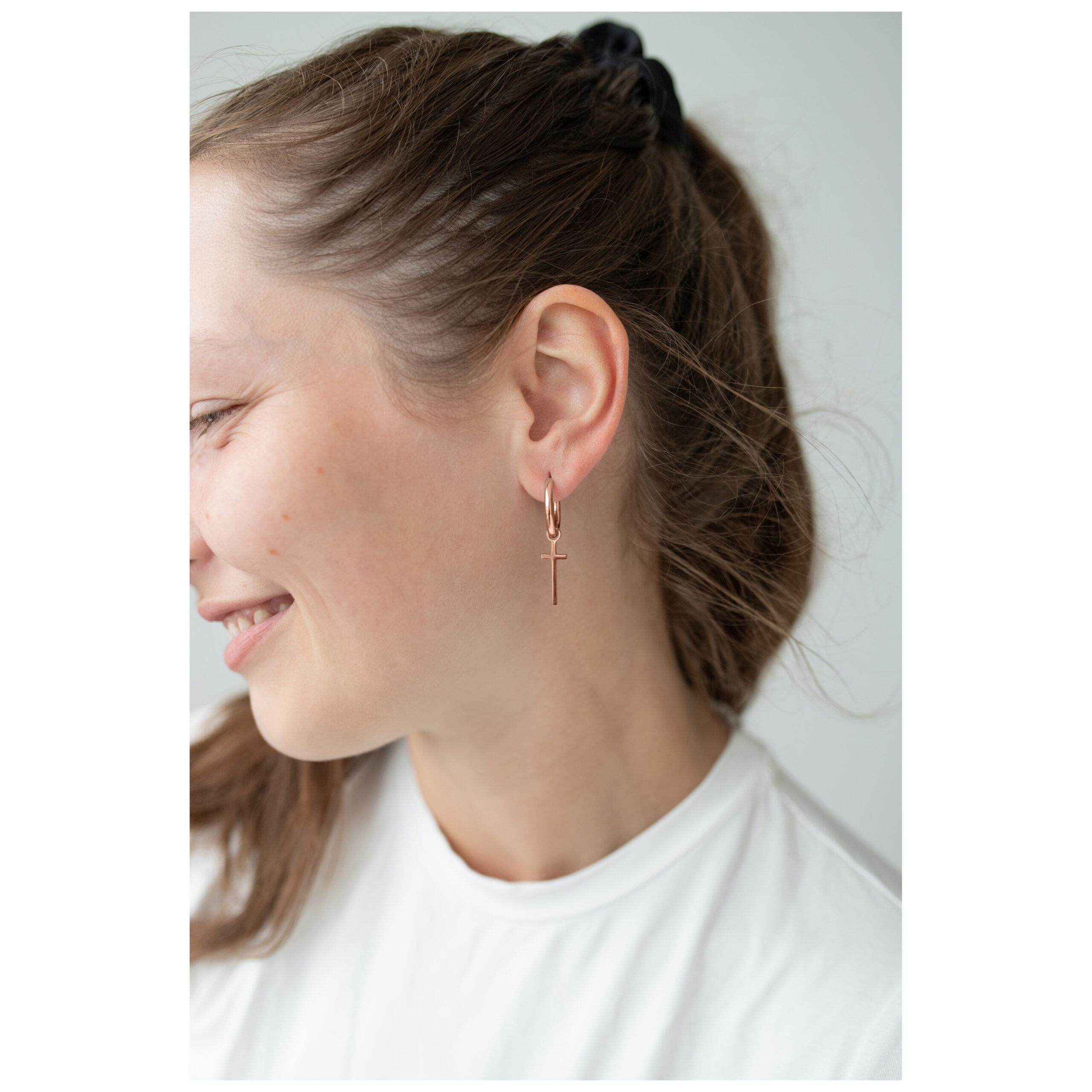 Roségoldene Little Cross Hoops im Ohr Roségoldene Creolen mit Kreuzanhänger am Ohr einer Frau, weißer Hintergrund