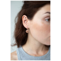 Frau trägt roségoldene Ohrringe mit Augenmotiv vor neutralem Hintergrund