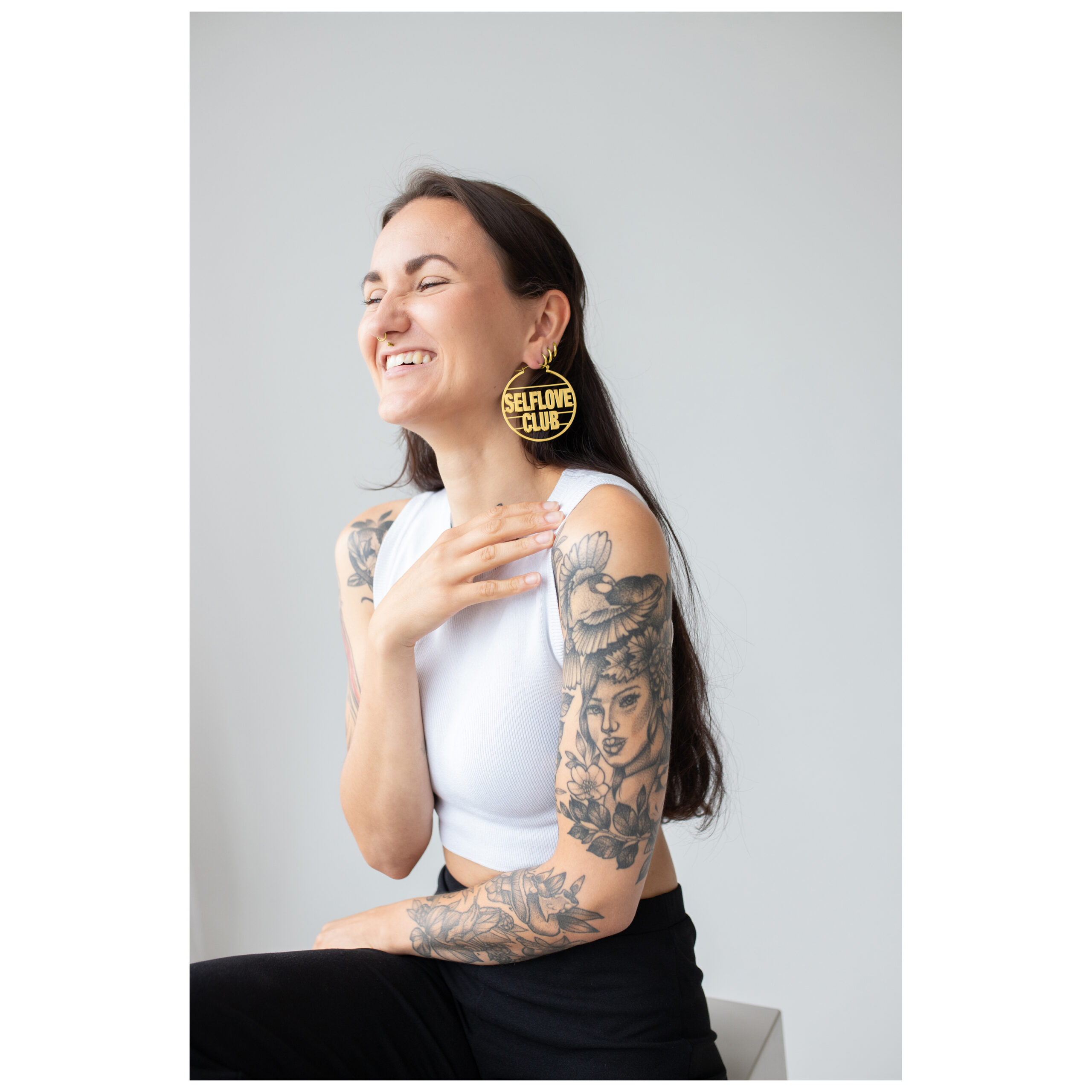 Mujer sonriente con aros Selflove Club dorados frente a un fondo neutro
