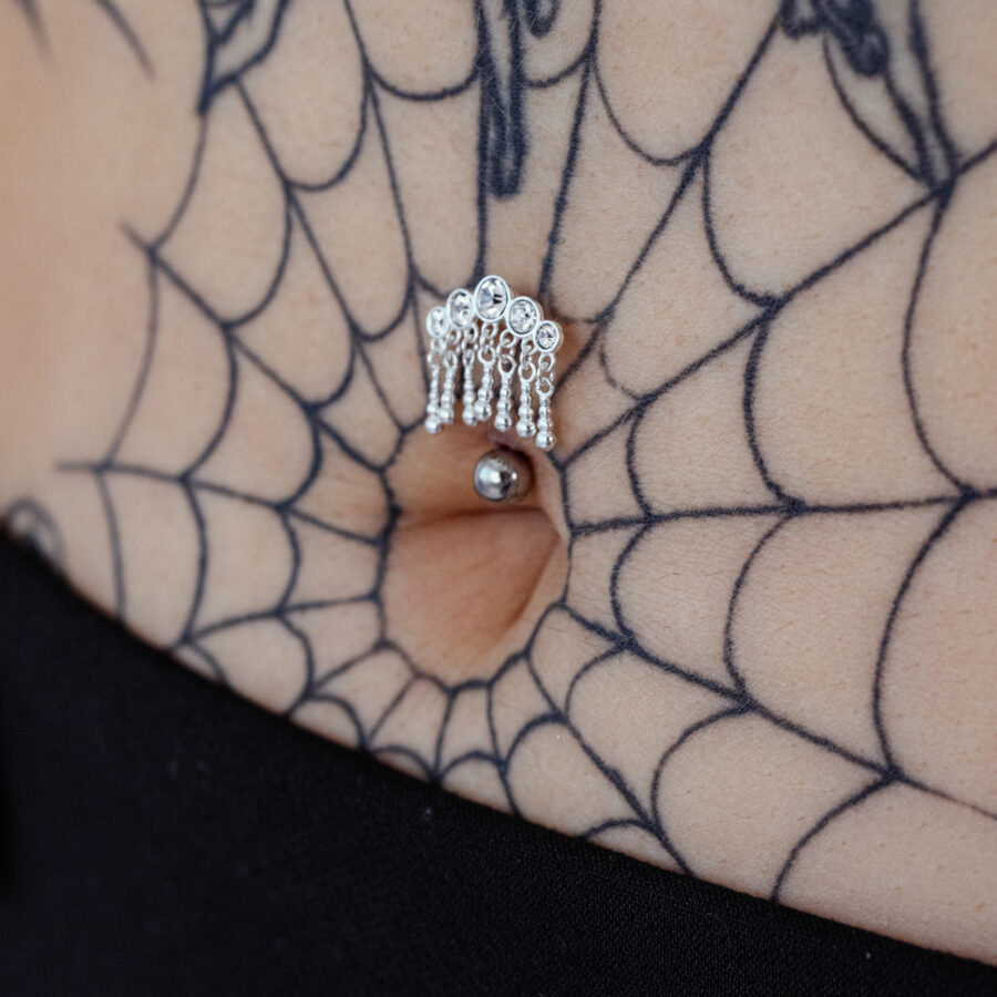 Piercing Bananabell de plata con cinco cristales colgantes en un ombligo, rodeado de un tatuaje de telaraña