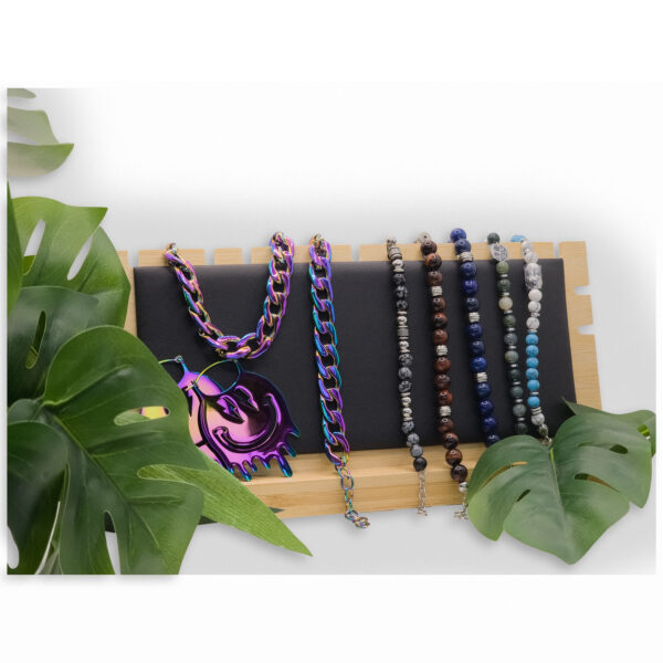 Expositor de cuero sintético negro con collares coloridos sobre madera, rodeado de hojas verdes