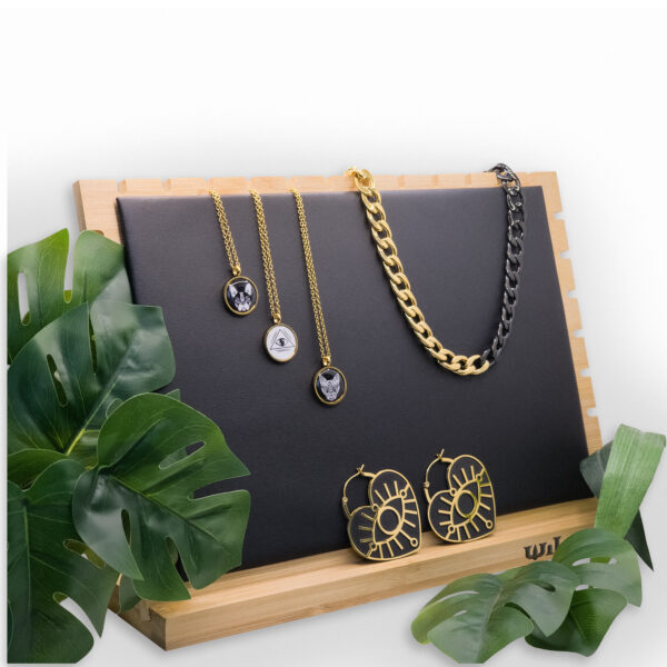 Expositor de joyas negro con cadenas doradas y pendientes sobre marco de madera, rodeado de hojas verdes