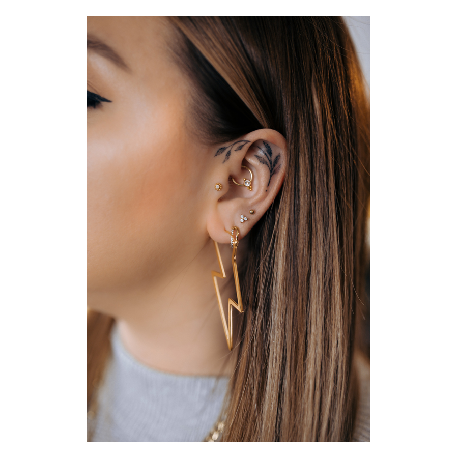 Aros Thunderbolt pendientes en oro Primer plano de una oreja con pendientes de rayo dorados y varios piercings, fondo neutro
