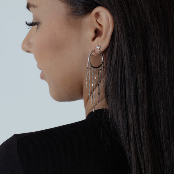 Mujer con cabello largo y oscuro lleva pendientes de borlas negras y plateadas sobre fondo blanco