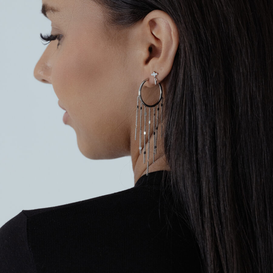 Mujer con cabello largo y oscuro lleva pendientes de borlas negras y plateadas sobre fondo blanco