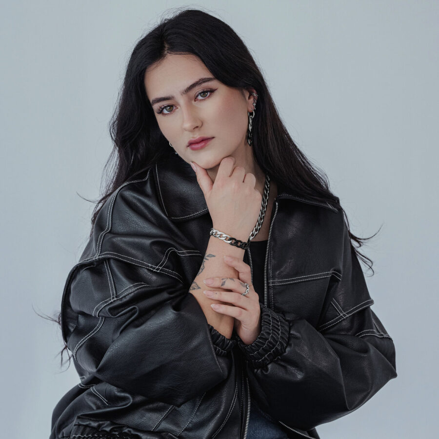 Mujer con chaqueta de cuero negra lleva un anillo de cadena plateado y una pulsera a juego sobre fondo gris