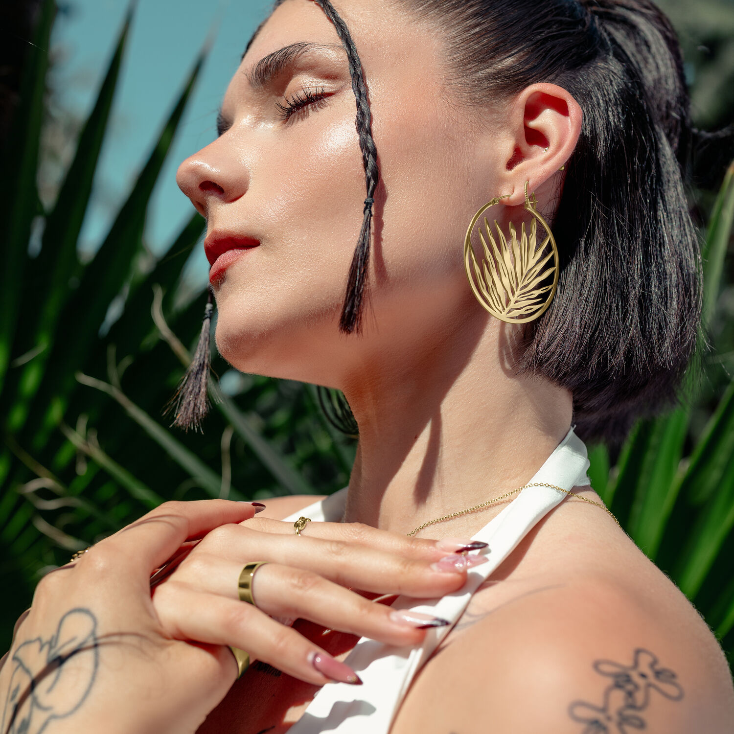 Aros Dorados Noble Leaf en la Oreja Mujer con pendientes dorados en forma de hoja, al aire libre frente a plantas verdes