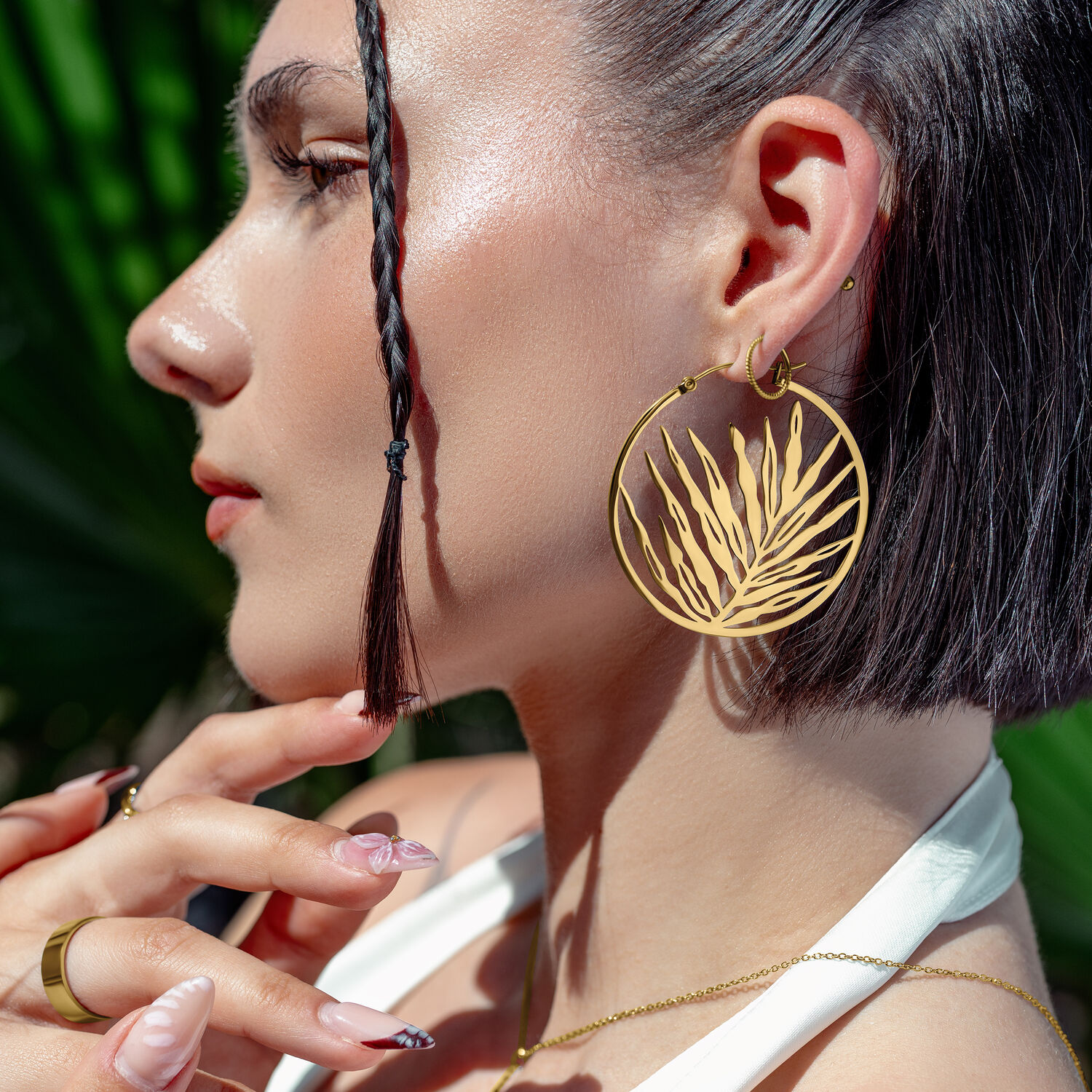 Aros dorados Noble Leaf en estilo veraniego Mujer con aros dorados Noble Leaf, de perfil frente a hojas verdes