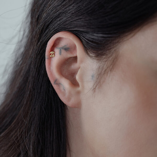 Accesorio Cherry dorado en la oreja, llevado por una persona con cabello oscuro, sobre fondo neutro