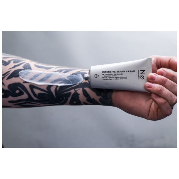 Mano sostiene No Cosmetics Crema Reparadora Intensiva, brazo con tatuaje al fondo
