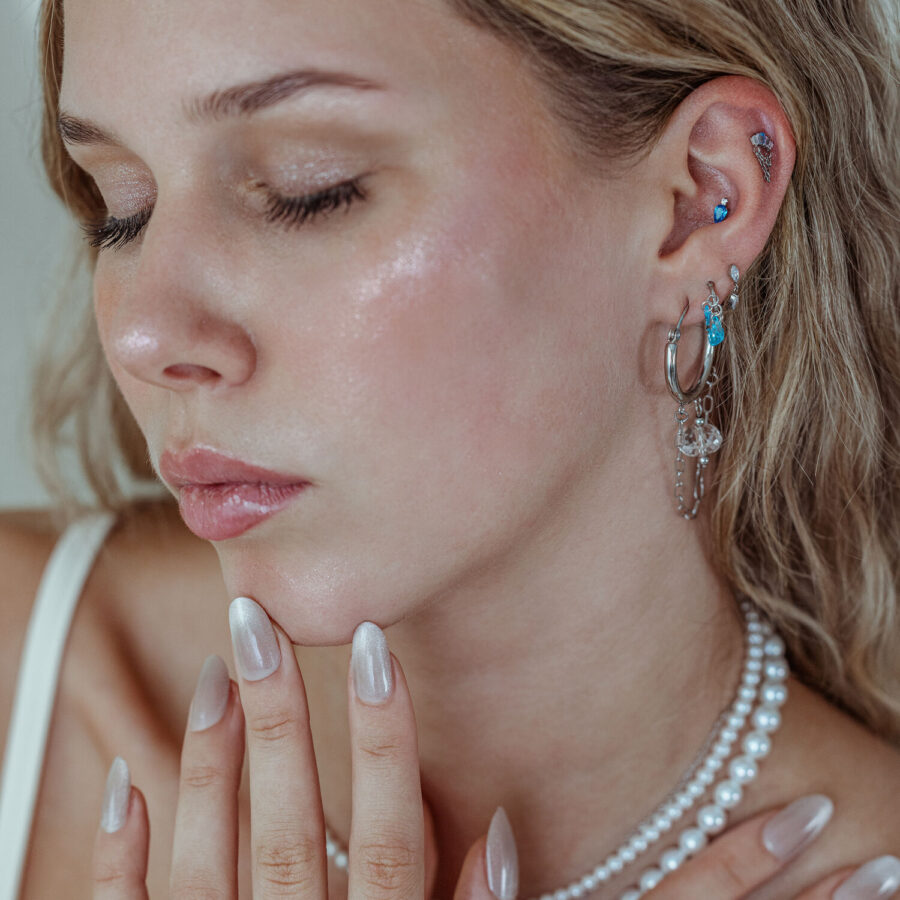 Mujer con varios piercings en la oreja, incluyendo el accesorio Mystic Dew en plata y oro con cristales lavanda y turquesa, sobre un fondo neutro