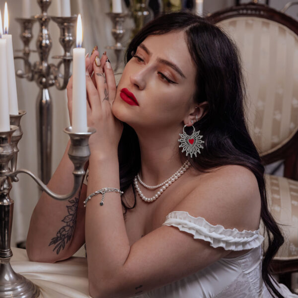 Mujer con cabello largo y oscuro lleva aros Sacred Heart en negro y plateado, sentada en una mesa con candelabro.