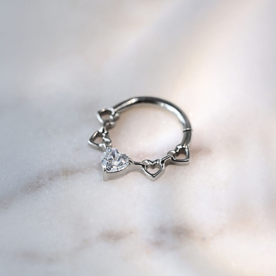 Clicker para septum plateado de titanio con diseño de corazones y cristales sobre fondo marmolado