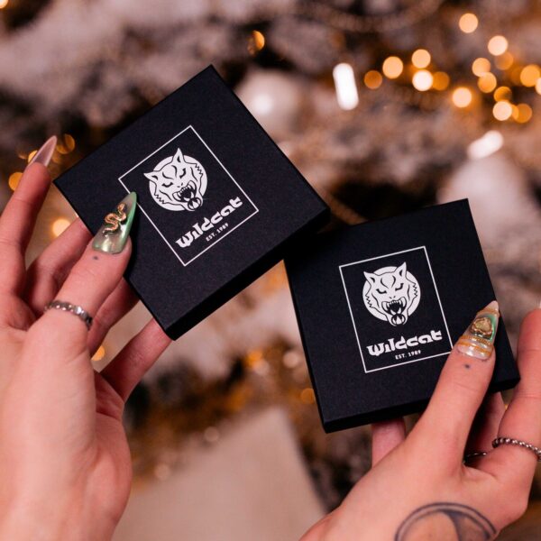 Dos cajas de regalo negras con el logo de Wildcat, sostenidas por manos con uñas doradas