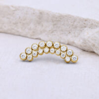 Accesorio de piercing dorado con cristales claros sobre fondo claro.
