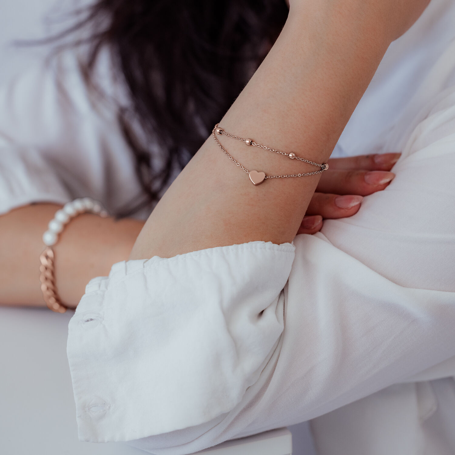 Pulsera de doble fila en oro rosa con colgante de corazón en la muñeca de una persona con camisa blanca