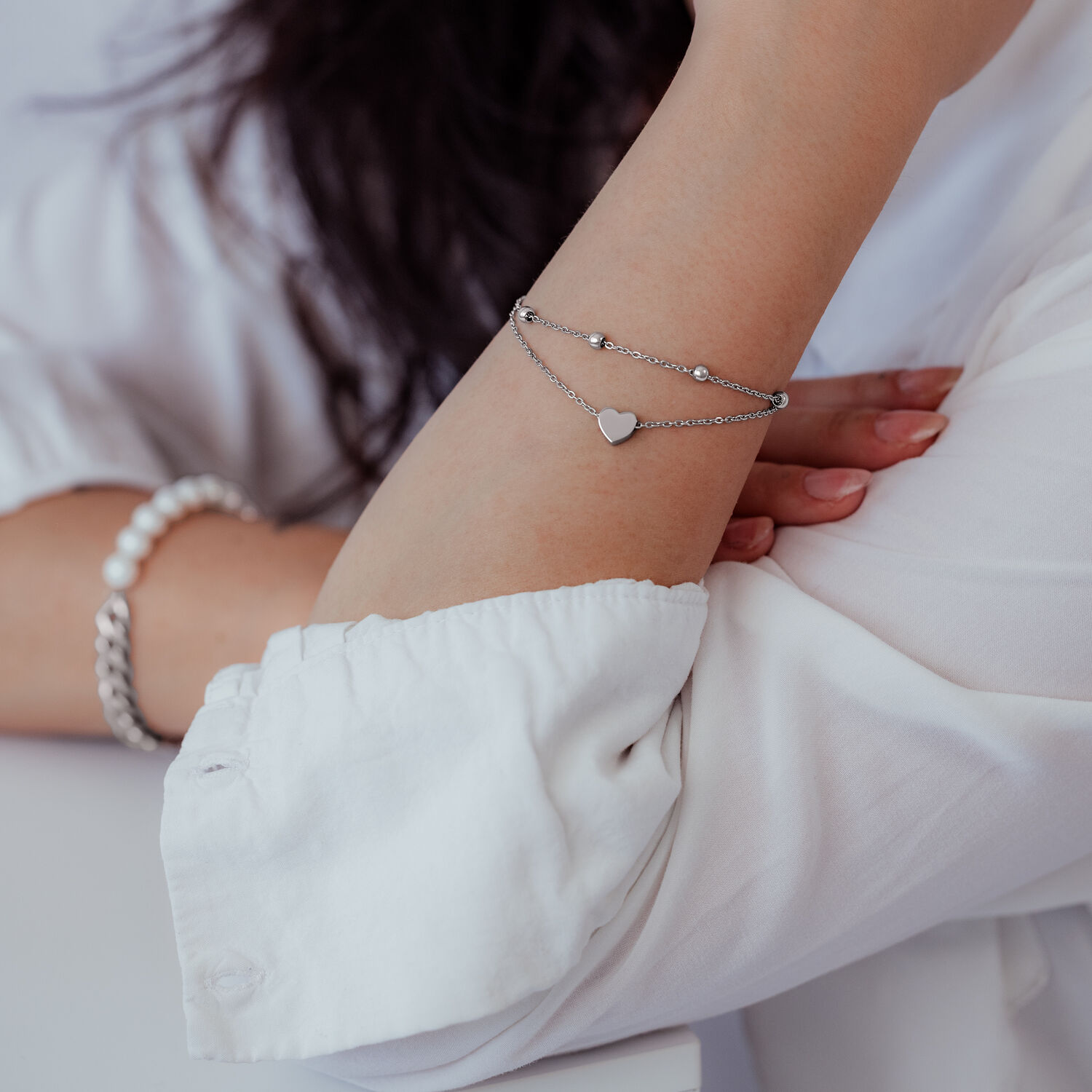 Pulsera de corazón de dos hileras en plata Pulsera plateada de dos hileras con colgante de corazón en el brazo de una persona con ropa blanca