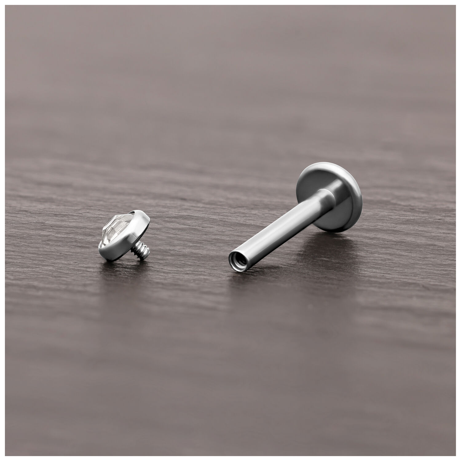 Cabezal de titanio plateado para piercing con cristal claro sobre madera oscura.