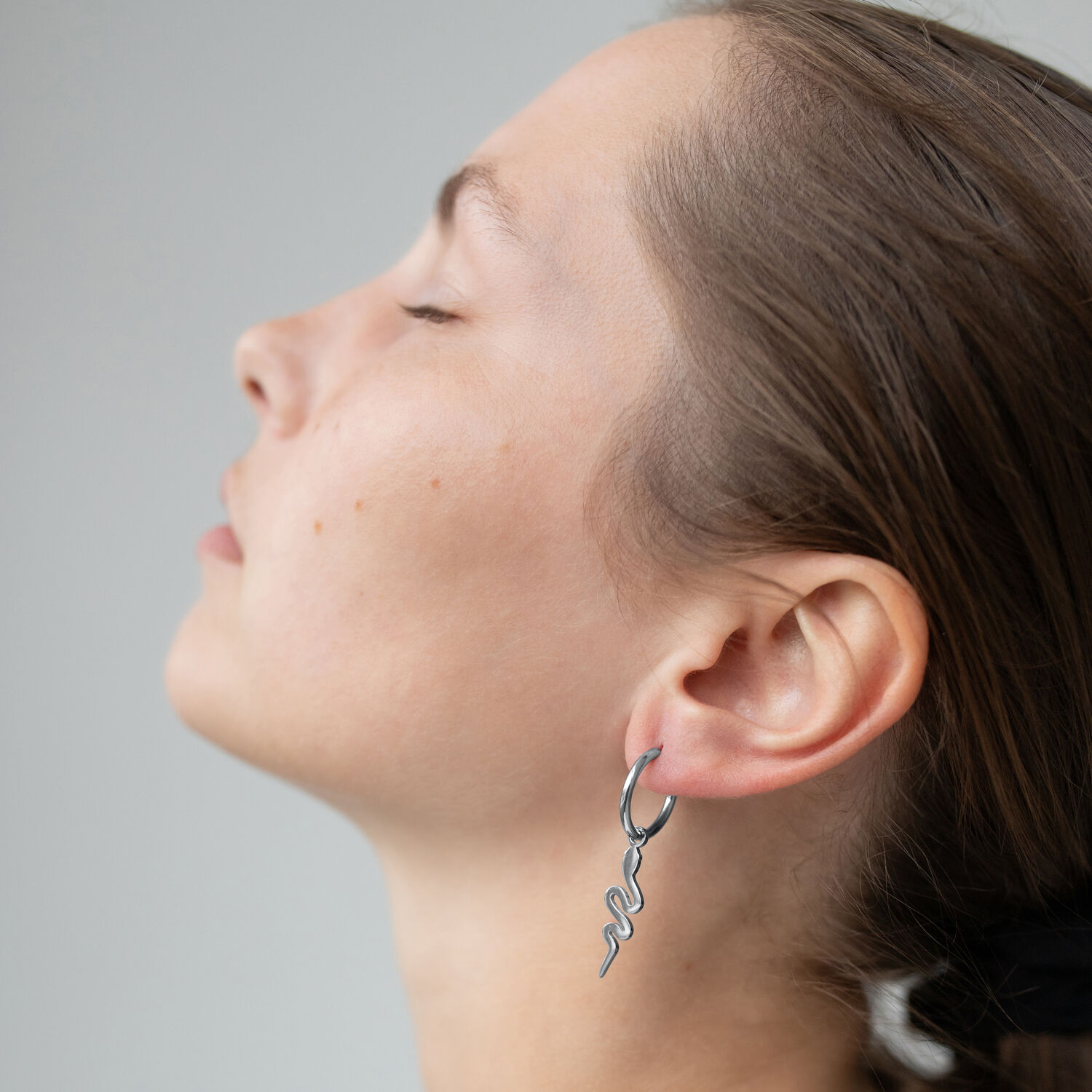 Perfil de una persona con los ojos cerrados que lleva pendientes de serpiente plateados, sobre fondo gris.