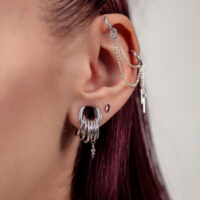 Primer plano de una oreja con cadena de piercing plateada y varios aros plateados