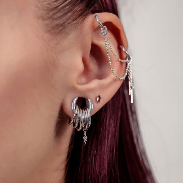 Primer plano de una oreja con cadena de piercing plateada y varios aros plateados