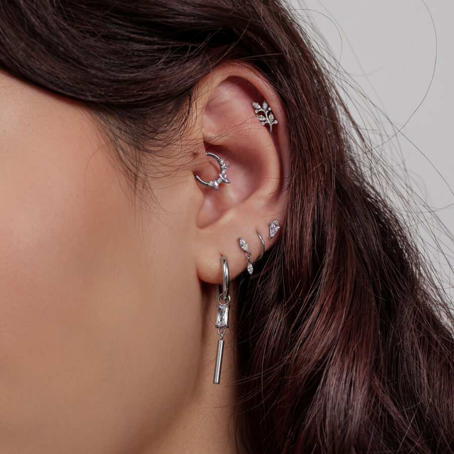 Oreja con varios piercings plateados, incluyendo diseños de hoja de cristal, sobre fondo claro
