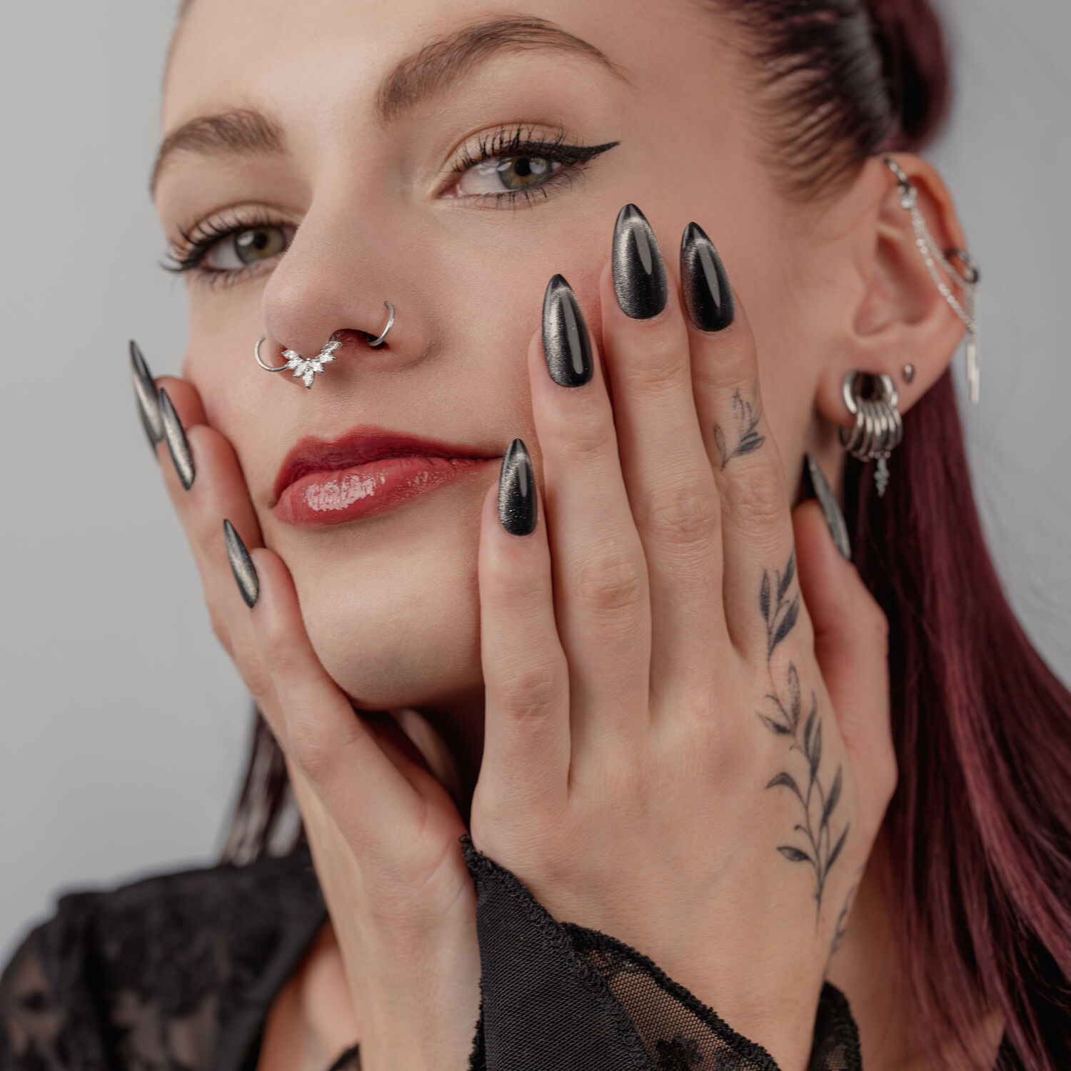Mujer con uñas negras brillantes, varios piercings en orejas y nariz, sobre fondo gris