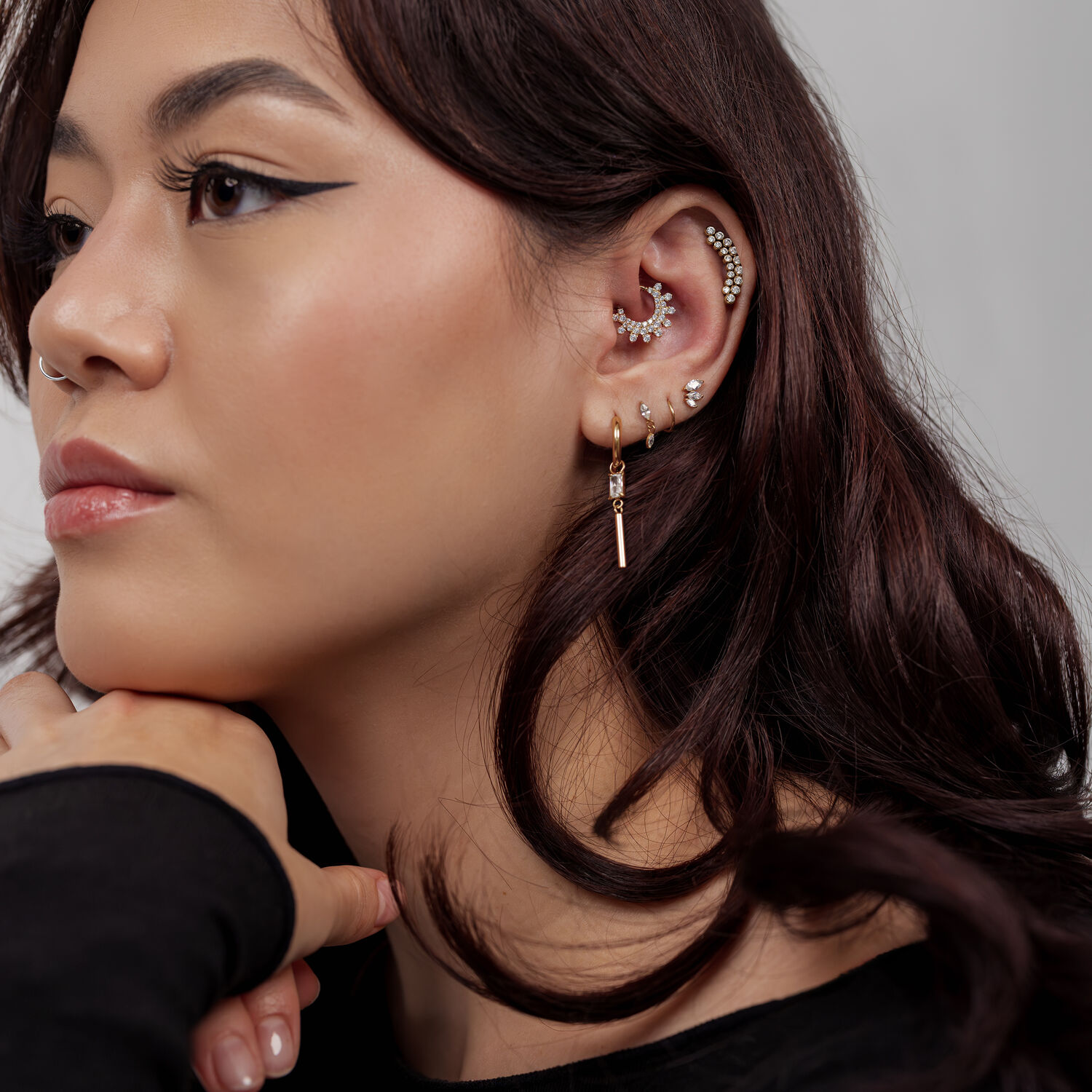 Mujer con varios piercings dorados en la oreja y un clicker de septum dorado sobre fondo gris