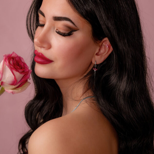 Mujer con cabello largo y oscuro sostiene una rosa rosa y lleva un pendiente de plata con cristal rojo sobre fondo rosa