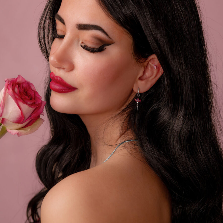Mujer con cabello largo y oscuro sostiene una rosa rosa y lleva un pendiente de plata con cristal rojo sobre fondo rosa