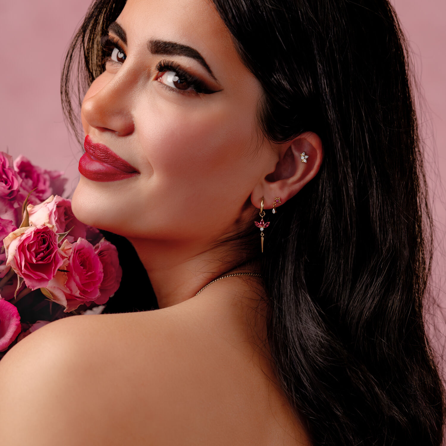Little Maharani Hoops con cristales rojos Mujer con cabello largo y negro lleva pendientes plateados con cristales rojos, sosteniendo rosas rosadas frente a un fondo rosa