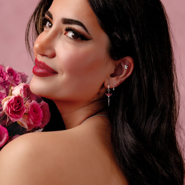 Mujer con cabello largo y oscuro lleva pendientes plateados con cristales rojos y sostiene rosas rosadas frente a un fondo rosa.