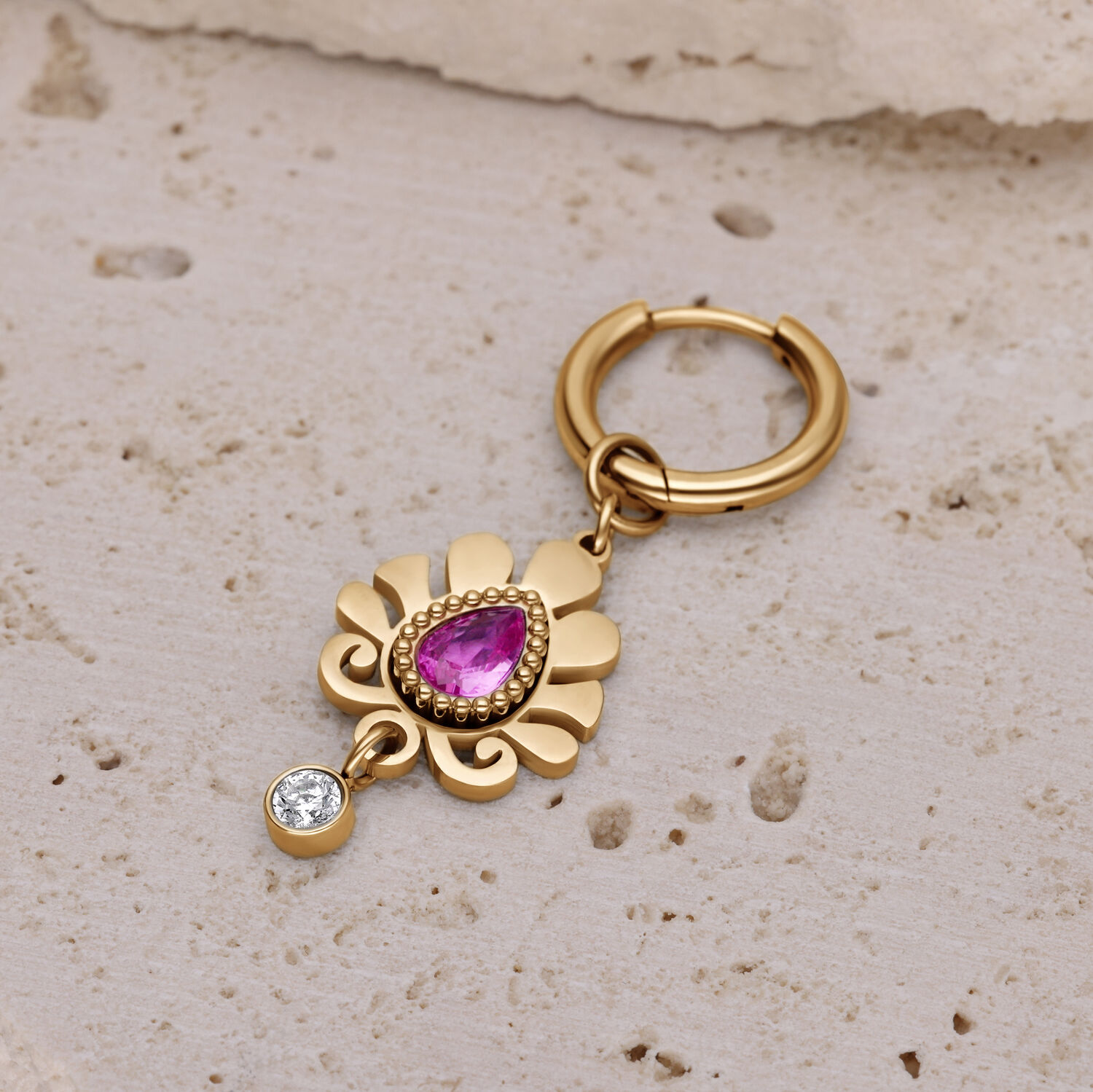 Pendiente dorado con colgante de cristal rosa Pendiente dorado con cristal rosa en forma de gota y pequeño colgante de cristal claro sobre superficie de piedra