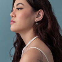 Mujer con cabello largo y castaño, pendientes de tortuga plateados, collar de perlas, fondo azul