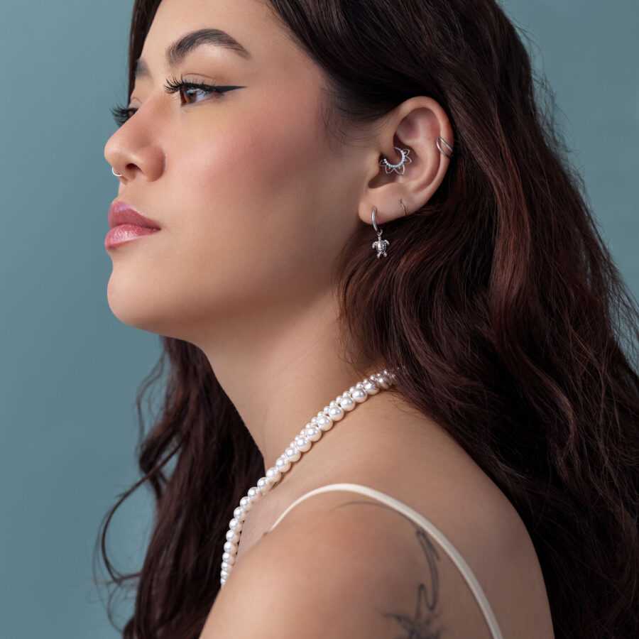 Mujer con cabello largo y castaño, pendientes de tortuga plateados, collar de perlas, fondo azul