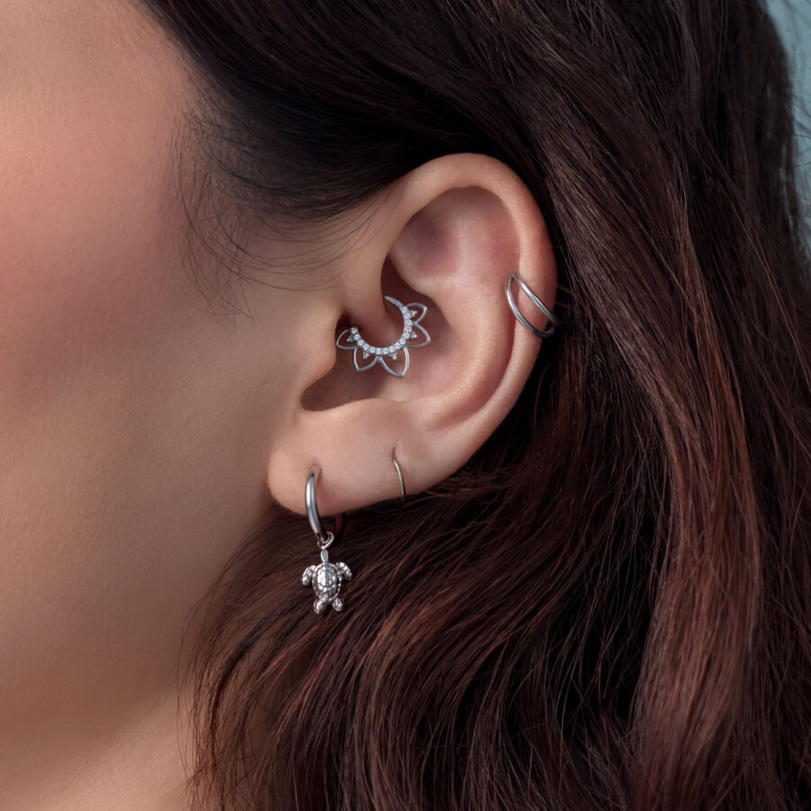 Primer plano de una oreja con pendientes de tortuga de plata y otros piercings, cabello castaño de fondo