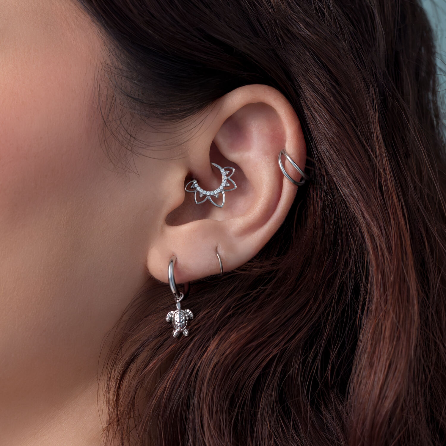 Primer plano de una oreja con pendientes de tortuga de plata y otros piercings, cabello castaño de fondo