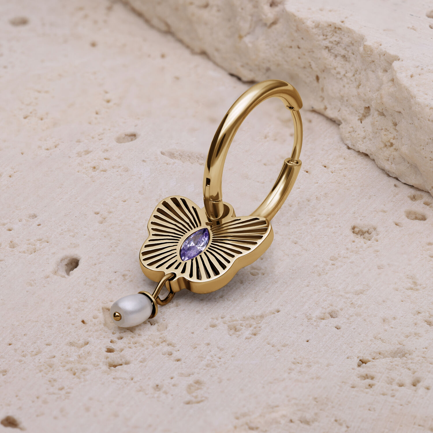 Pendiente dorado con piedra lavanda y perla Pendiente dorado con diseño floral, piedra lavanda y colgante de perla sobre fondo de piedra clara