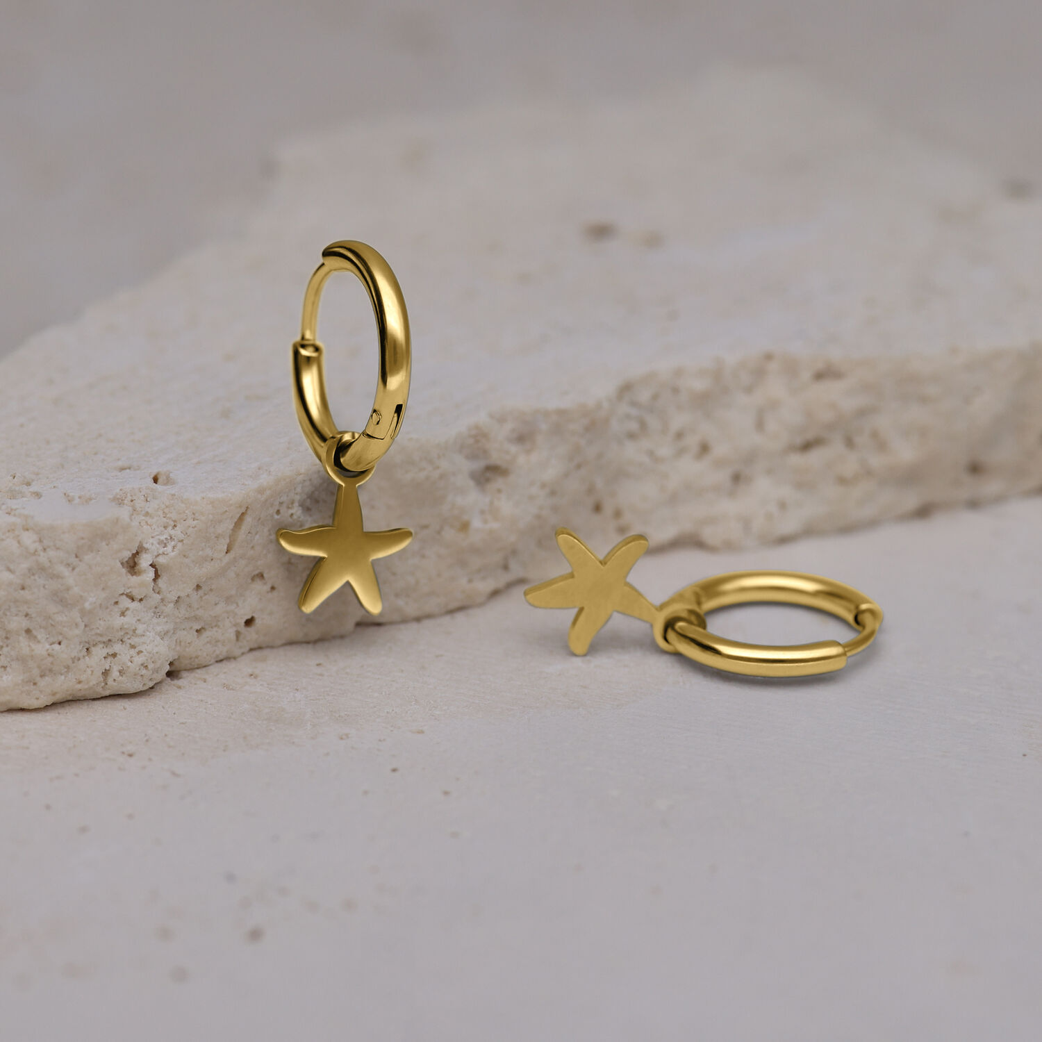 Mini aros dorados con estrella de mar Zlata Dos aros dorados con colgantes de estrella de mar sobre una superficie de piedra