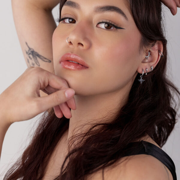 Mujer con cabello largo lleva pendientes Mini Branch de acero quirúrgico, varios piercings y tatuajes visibles.