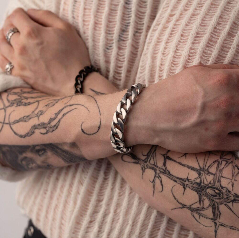 Persona con brazos tatuados lleva un brazalete chunky plateado, brazos cruzados sobre un suéter color crema