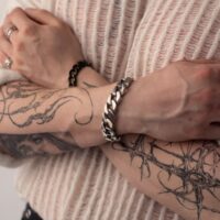 Persona con brazos tatuados lleva un brazalete chunky plateado, brazos cruzados sobre un suéter color crema