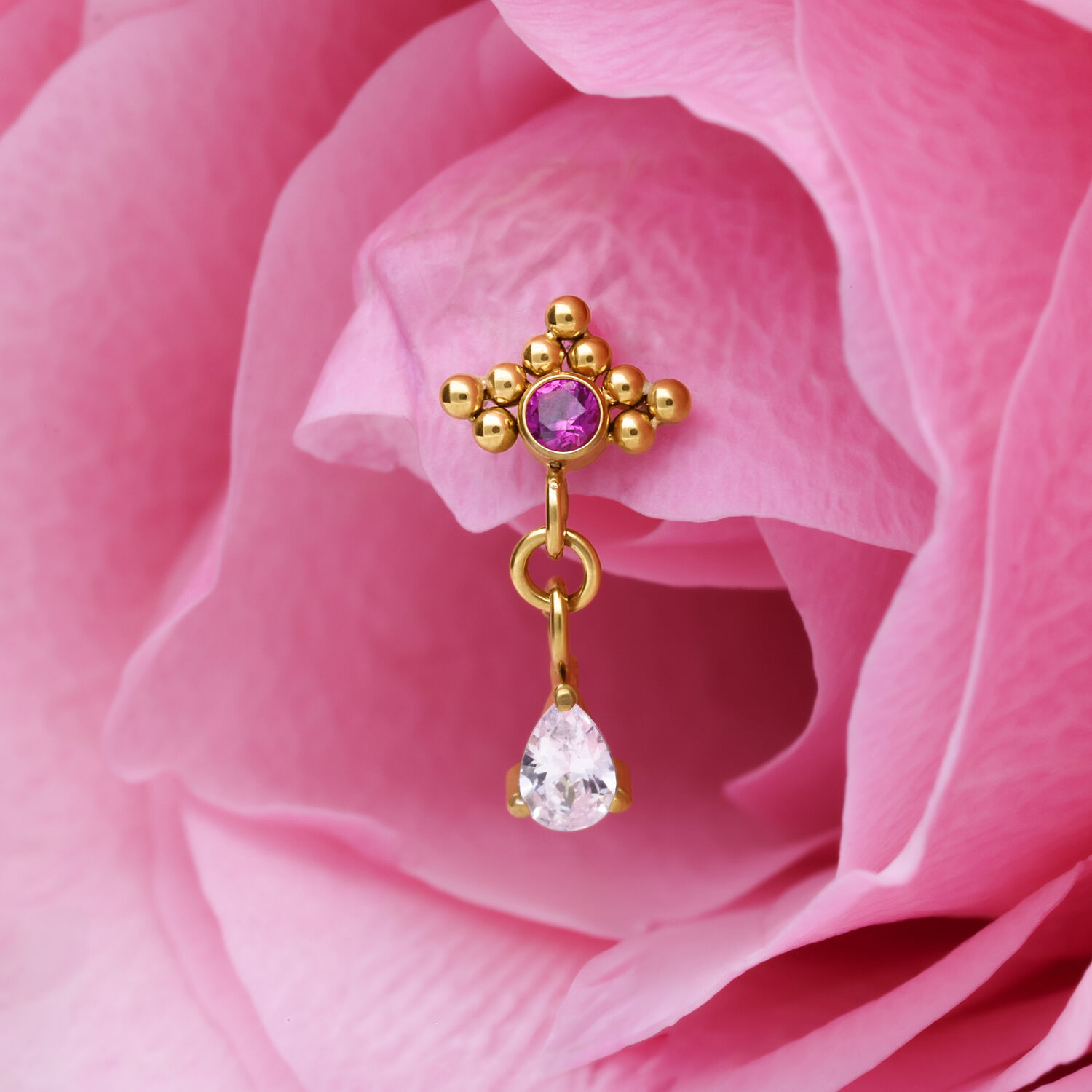 Pieza de piercing dorada con cristal rojo y colgante en forma de gota sobre pétalo rosa