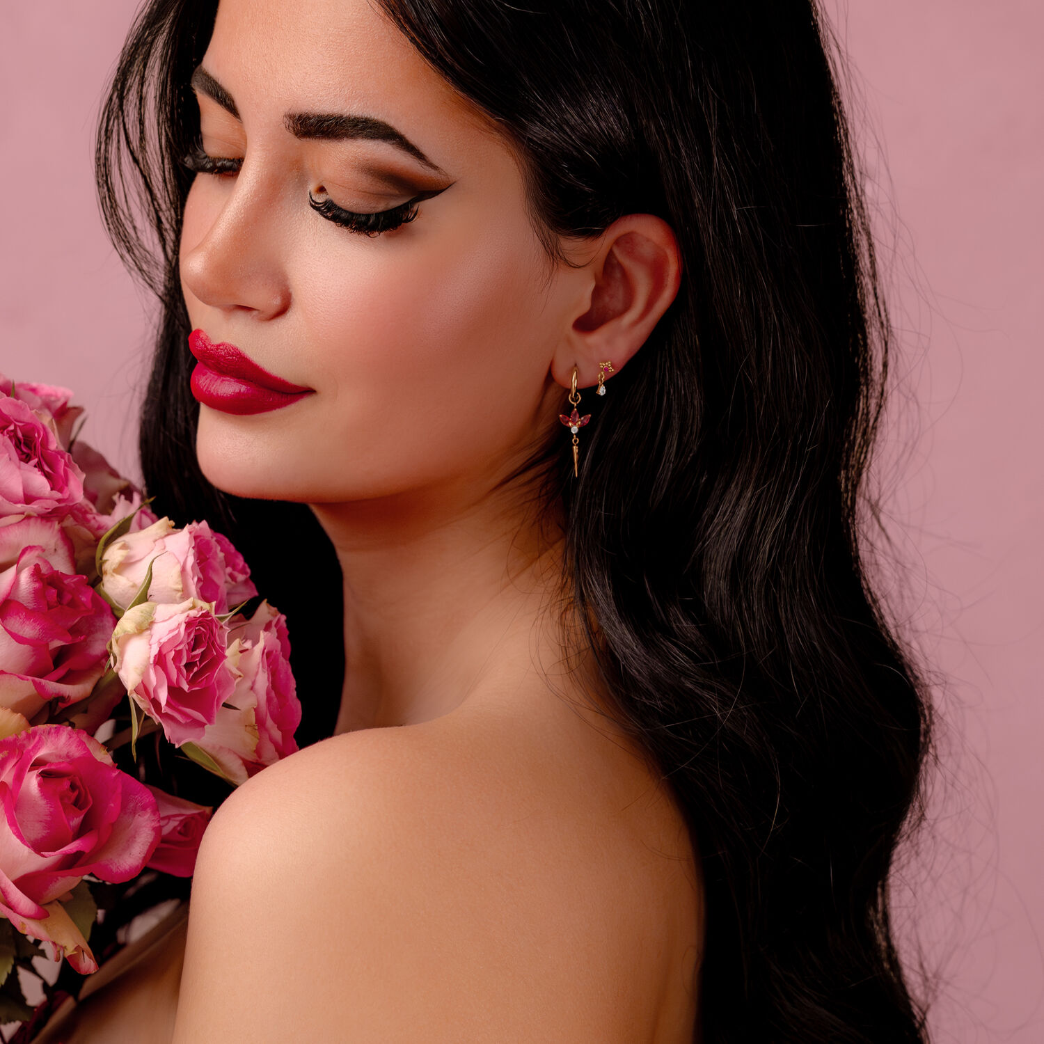 Mujer con cabello largo y negro, labios rojos y pendientes dorados, sosteniendo rosas rosadas sobre fondo rosa