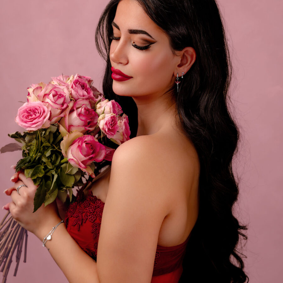 Mujer con cabello largo y oscuro sostiene un ramo de rosas rosadas frente a un fondo rosa.