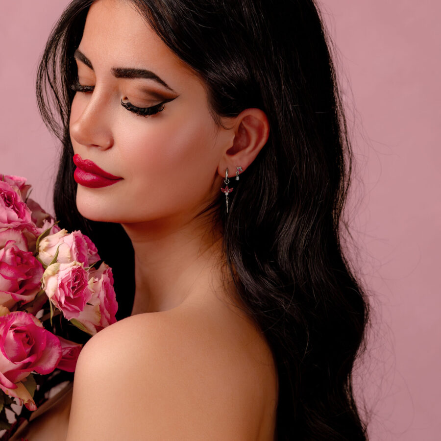Mujer con cabello largo y oscuro y pendientes de plata con cristales rojos, sosteniendo flores rosas sobre un fondo rosa