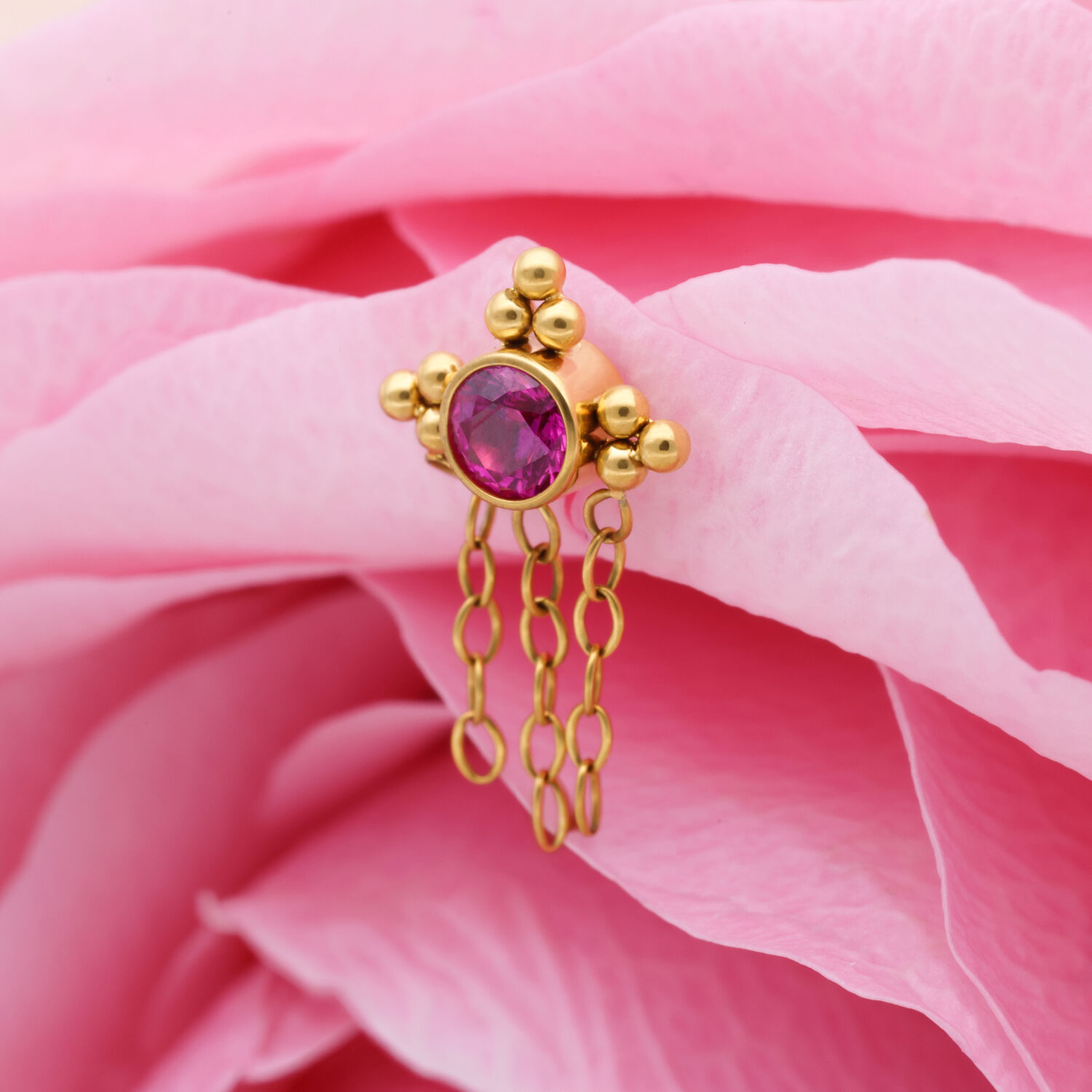 Accesorio de titanio dorado con cristal rojo y cadenas, sobre pétalo rosa